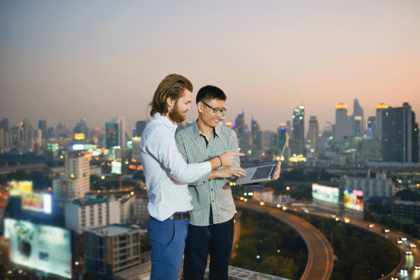 businessmen-using-laptop-while-standing-against-cityscape-sunset-scaled